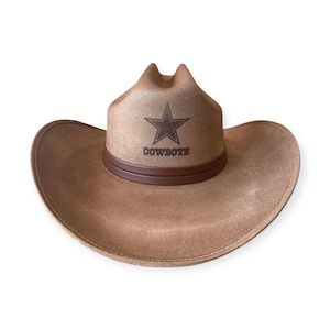 Puede incluir: Un sombrero de vaquero marr&oacute;n claro con una banda de cuero marr&oacute;n. El sombrero presenta una estrella marr&oacute;n oscuro y la palabra "COWBOYS" impresa en la parte delantera. El sombrero tiene un dise&ntilde;o occidental cl&aacute;sico.