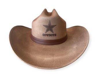 May include: A tan cowboy hat with a brown leather band. The hat features a dark brown star and the word "COWBOYS" printed on the front. The hat has a classic western design.