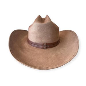 Puede incluir: Un sombrero de vaquero color canela con ala ancha y una banda de cuero marr&oacute;n. El sombrero tiene un dise&ntilde;o cl&aacute;sico del oeste, perfecto para actividades al aire libre o eventos de disfraces. El sombrero es de material suave.
