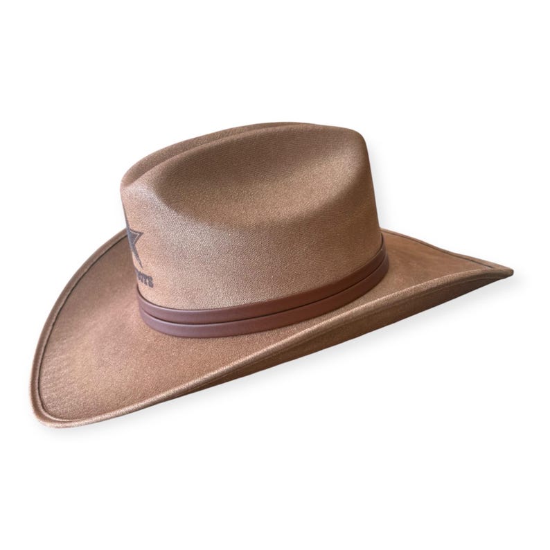 Puede incluir: Sombrero de vaquero color canela con una banda de cuero marr&oacute;n y un logotipo de estrella. El sombrero tiene un ala ancha y una corona redondeada. El sombrero est&aacute; hecho de un material suave similar al ante.