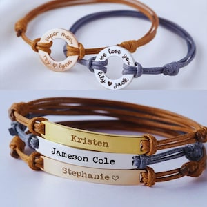 Op de afbeelding: Twee armbanden met gepersonaliseerde bedels. De bovenste armband heeft een zilveren ronde bedel met de woorden "we love you" en "elly jack" erop gegraveerd. De onderste armband heeft drie rechthoekige bedels van goud en zilver met de namen "Kristen", "Jameson Cole" en "Stephanie" erop gegraveerd.
