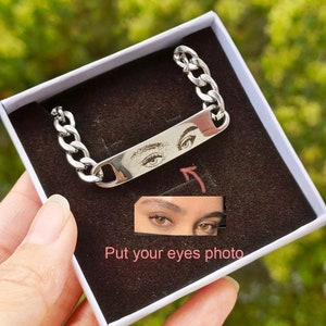 May include: A silver chain bracelet with a rectangular tag engraved with a pair of eyes. The tag is attached to the chain with a silver clasp. The bracelet is in a white box with a black interior. The text "Put your eyes photo" is written below the bracelet.