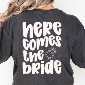 Back of a dark t-shirt reading ‘here comes the bride’ in Jelly Puff cute chunky handwritten font, soft blush ink, stacked lines with a small diamond ring doodle. Playful all-caps mix-case display for bridal party shirts.