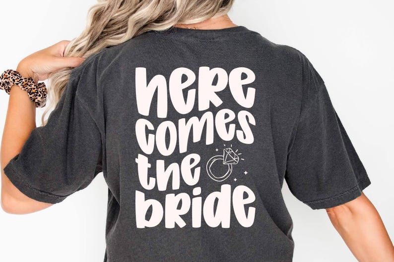 Back of a dark t-shirt reading ‘here comes the bride’ in Jelly Puff cute chunky handwritten font, soft blush ink, stacked lines with a small diamond ring doodle. Playful all-caps mix-case display for bridal party shirts.