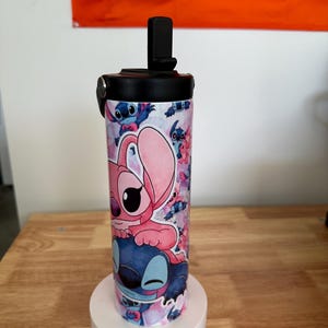 May include: A stainless steel water bottle featuring a colorful design with the characters Stitch and Angel from the animated movie Lilo & Stitch. The bottle has a black lid with a flip-top opening and is approximately 8 inches tall.