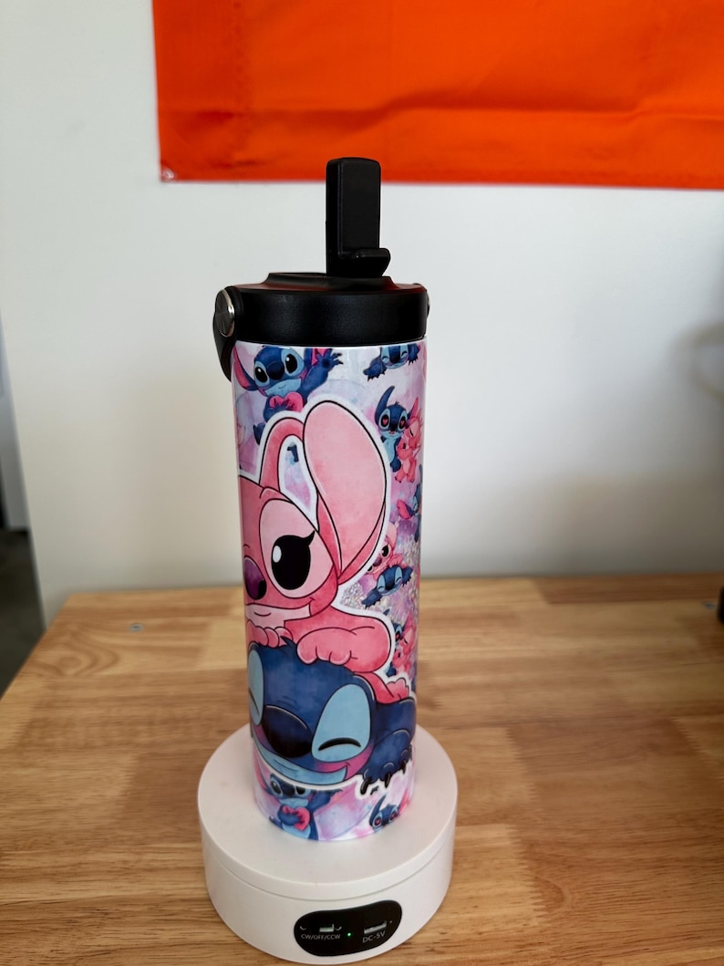 May include: A stainless steel water bottle featuring a colorful design with the characters Stitch and Angel from the animated movie Lilo & Stitch. The bottle has a black lid with a flip-top opening and is approximately 8 inches tall.