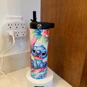 Puede incluir: Una base de carga blanca con una botella de agua de acero inoxidable azul y rosa con un personaje de dibujos animados de Stitch de Lilo & Stitch. La botella tiene una tapa negra con una pajita.