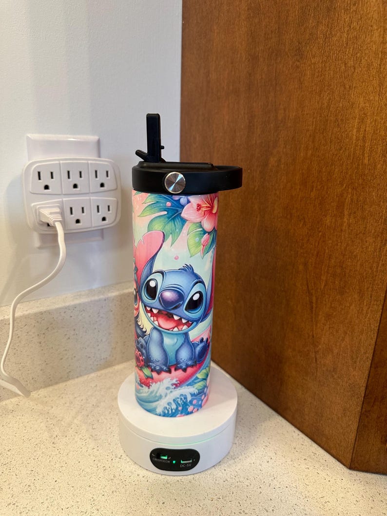Puede incluir: Una base de carga blanca con una botella de agua de acero inoxidable azul y rosa con un personaje de dibujos animados de Stitch de Lilo & Stitch. La botella tiene una tapa negra con una pajita.