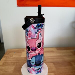 May include: A stainless steel water bottle featuring a colorful design with the characters Stitch and Angel from the animated film Lilo & Stitch. The bottle has a black lid with a handle and a flip-top straw. The background is white and orange.