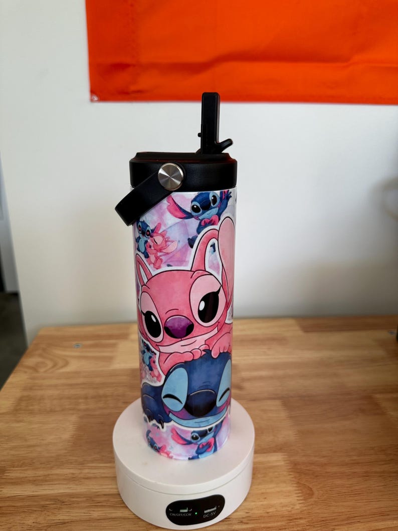 May include: A stainless steel water bottle featuring a colorful design with the characters Stitch and Angel from the animated film Lilo & Stitch. The bottle has a black lid with a handle and a flip-top straw. The background is white and orange.