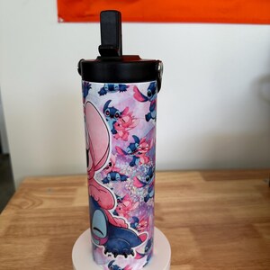May include: A cylindrical tumbler featuring a colorful design of the character Stitch from the animated film Lilo & Stitch. The tumbler has a black lid with a flip-top opening and a black handle. The background of the design is pink and purple.