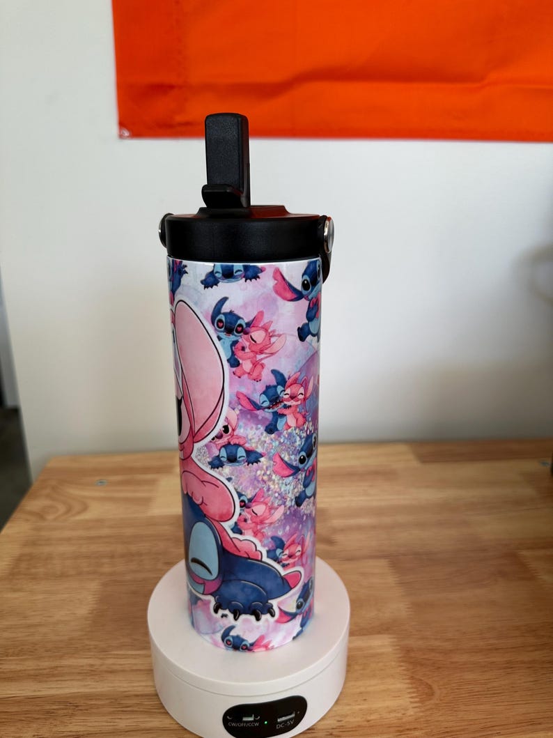 May include: A cylindrical tumbler featuring a colorful design of the character Stitch from the animated film Lilo & Stitch. The tumbler has a black lid with a flip-top opening and a black handle. The background of the design is pink and purple.