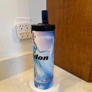 May include: A white and blue insulated water bottle with a black lid and a picture of Elsa from Frozen. The bottle is on a white charging base with a green light.