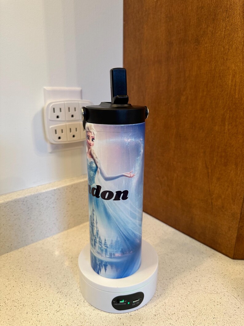 May include: A white and blue insulated water bottle with a black lid and a picture of Elsa from Frozen. The bottle is on a white charging base with a green light.