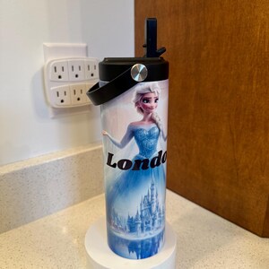 May include: A white and black insulated tumbler with a black lid and handle. The tumbler features a colorful image of a princess character from the movie Frozen, with the name "London" printed on the side. The tumbler is sitting on a white charging base.