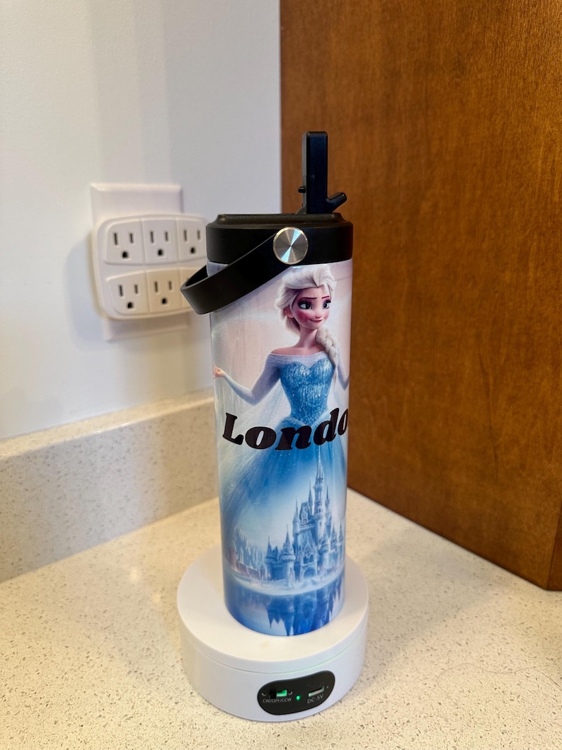 May include: A white and black insulated tumbler with a black lid and handle. The tumbler features a colorful image of a princess character from the movie Frozen, with the name "London" printed on the side. The tumbler is sitting on a white charging base.