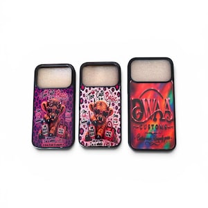 May include: Three phone cases with black borders. Two cases feature a cartoon Highland cow wearing sunglasses and holding a purse, with the text "Too Cute to be Basic". The third case has a red and orange gradient with "WAA CUSTOMS" text.