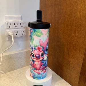 Puede incluir: Una base de carga blanca con una botella de agua negra y azul con un dise&ntilde;o de personaje de dibujos animados rosa y azul. La botella tiene una tapa negra con una manija negra. La base de carga tiene una luz verde. El texto en la base de carga dice "DC 5V".