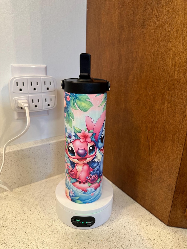 Puede incluir: Una base de carga blanca con una botella de agua negra y azul con un dise&ntilde;o de personaje de dibujos animados rosa y azul. La botella tiene una tapa negra con una manija negra. La base de carga tiene una luz verde. El texto en la base de carga dice "DC 5V".