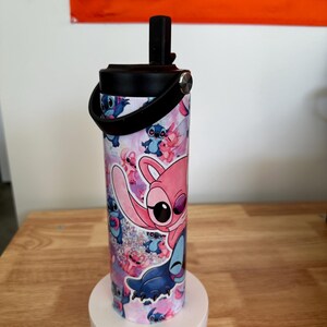 May include: A cylindrical water bottle featuring a colorful design of the character Stitch from the animated film Lilo & Stitch. The bottle has a black lid with a handle and is approximately 8 inches tall. The design includes pink, blue, and purple hues.