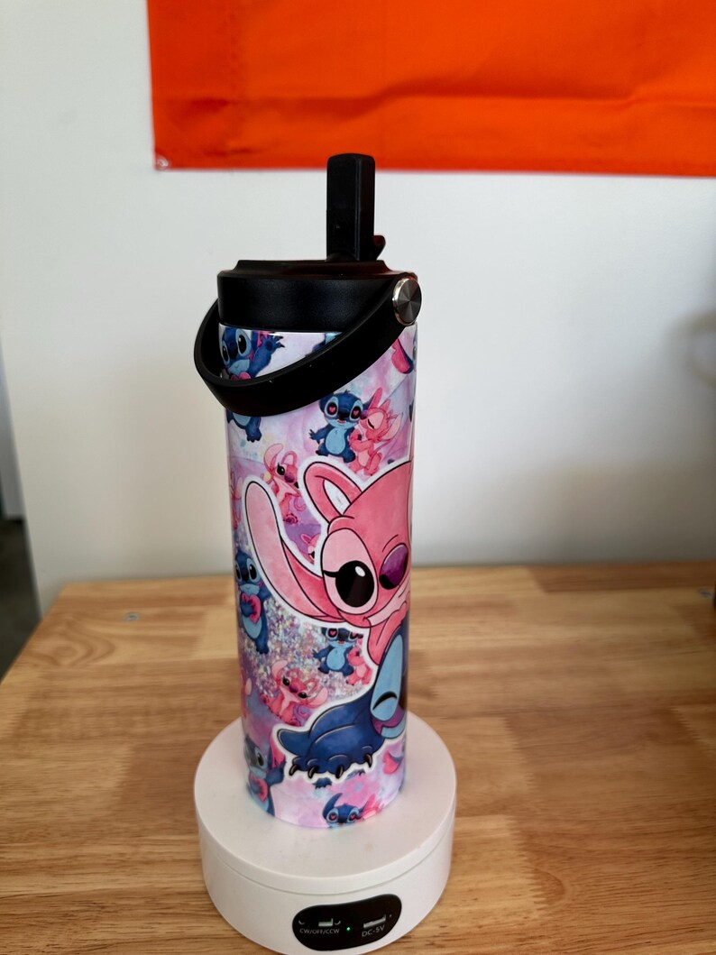 May include: A cylindrical water bottle featuring a colorful design of the character Stitch from the animated film Lilo & Stitch. The bottle has a black lid with a handle and is approximately 8 inches tall. The design includes pink, blue, and purple hues.
