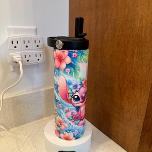 Puede incluir: Una base de carga blanca con un vaso estampado en rosa y azul con un personaje de dibujos animados. El vaso tiene una tapa negra y una pajita negra.