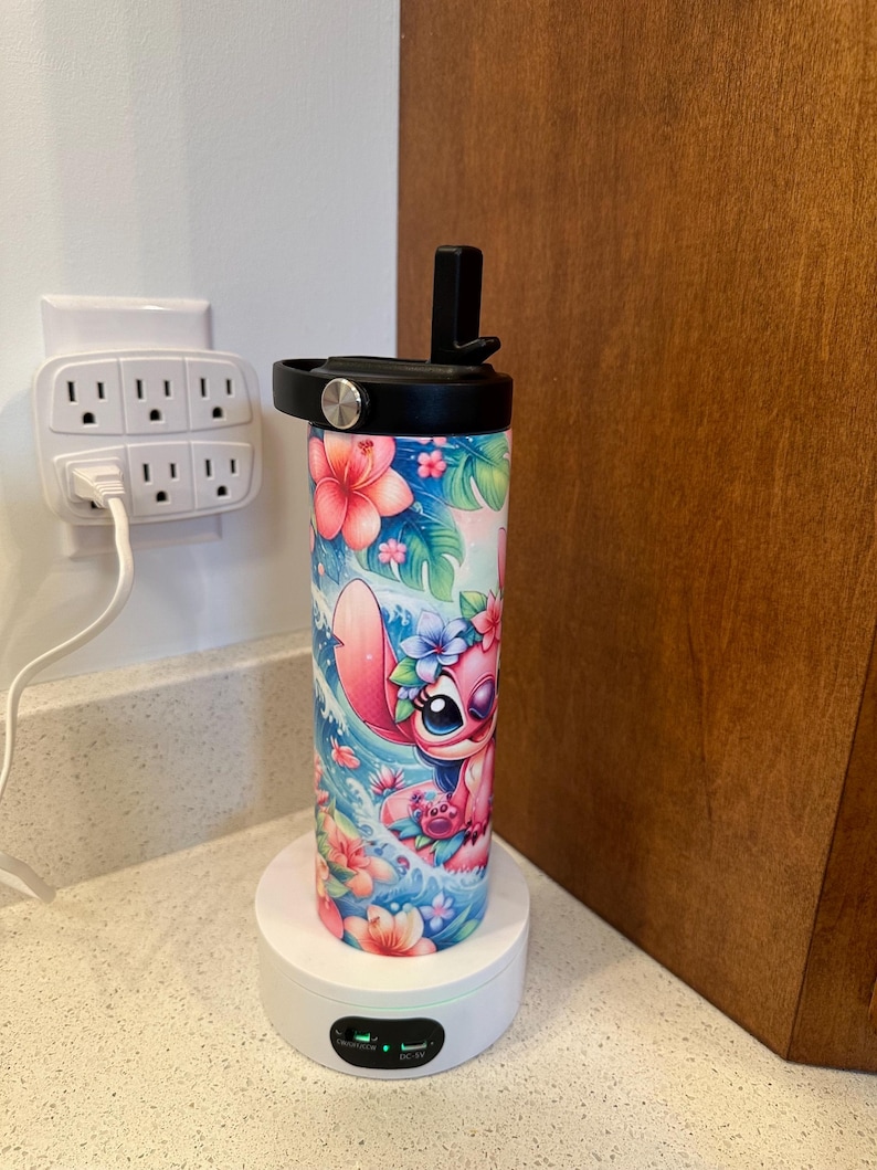 Puede incluir: Una base de carga blanca con un vaso estampado en rosa y azul con un personaje de dibujos animados. El vaso tiene una tapa negra y una pajita negra.