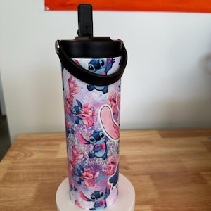 May include: A stainless steel tumbler featuring a colorful pattern of the character Stitch from the animated film Lilo & Stitch. The tumbler has a black lid with a handle and a flip-top opening. The design includes Stitch in various poses against a purple and pink background.
