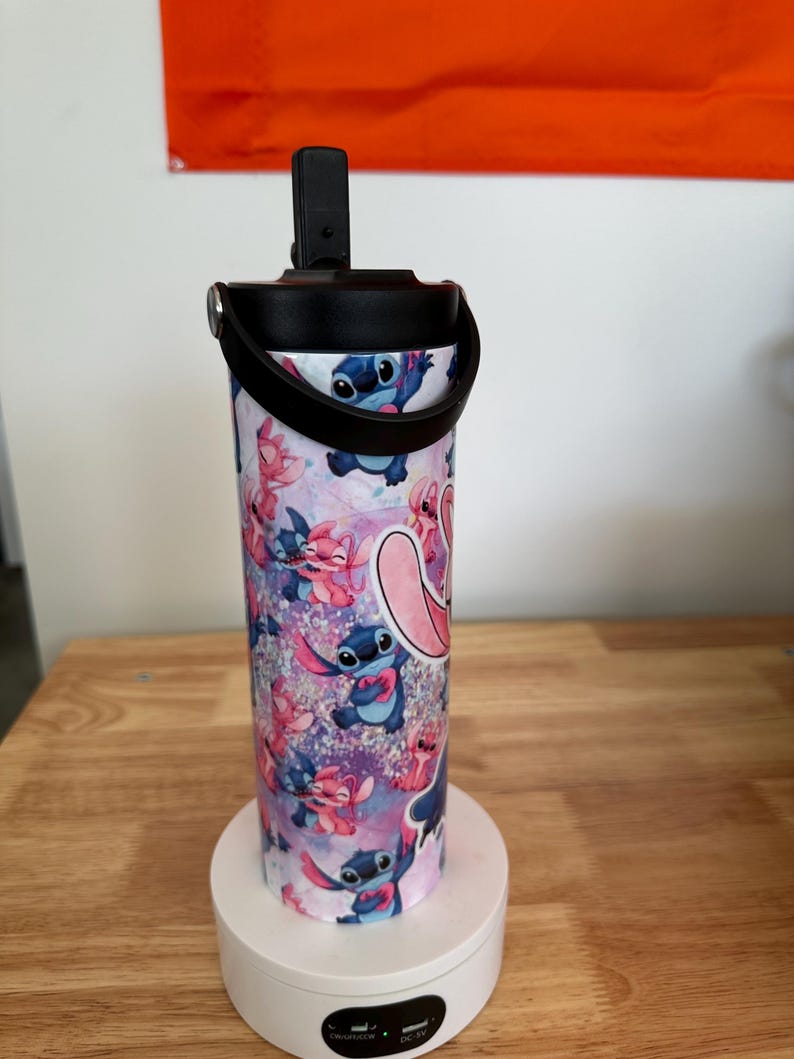 May include: A stainless steel tumbler featuring a colorful pattern of the character Stitch from the animated film Lilo & Stitch. The tumbler has a black lid with a handle and a flip-top opening. The design includes Stitch in various poses against a purple and pink background.