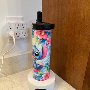 Puede incluir: Una base de carga blanca con un indicador de luz verde y un puerto de carga negro. Una botella de agua azul y rosa con un dise&ntilde;o floral y un personaje de dibujos animados de Stitch de Lilo & Stitch est&aacute; en la base de carga.