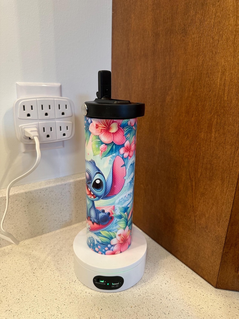 Puede incluir: Una base de carga blanca con un indicador de luz verde y un puerto de carga negro. Una botella de agua azul y rosa con un dise&ntilde;o floral y un personaje de dibujos animados de Stitch de Lilo & Stitch est&aacute; en la base de carga.