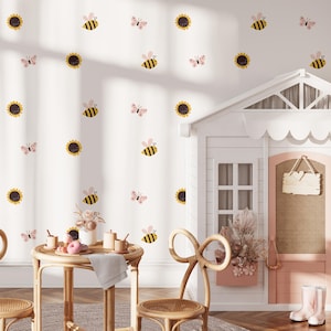 Könnte beinhalten: Eine weiße Wand mit Aufklebern von Bienen, Schmetterlingen und Sonnenblumen. Ein kleiner Tisch und Stühle stehen vor der Wand. Ein Spielhaus mit rosa Tür und Schild befindet sich auf der rechten Seite.