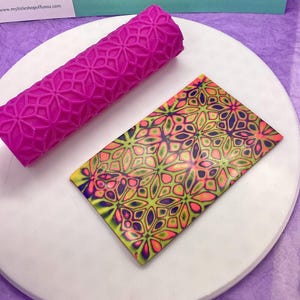 May include: A pink silicone rolling pin with a geometric pattern, used to create a decorative design on a sheet of colourful polymer clay. The clay is a vibrant mix of green, yellow, pink, and purple.