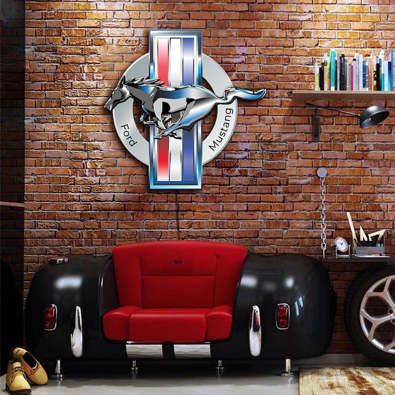 May include: A red leather couch shaped like a classic car with a chrome trim. The couch is in front of a brick wall with a Ford Mustang logo on it. The logo is silver and blue with the words "Ford" and "Mustang" on it.