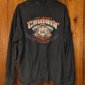 May include: Black long-sleeve t-shirt with a graphic print of a motorcycle engine and the text "St. Louis Cruisin' On Route 66 Docs Harley-Davidson Kirkwood, Missouri".