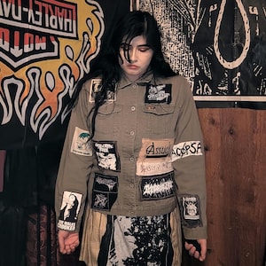May include: Olive green denim jacket adorned with multiple patches featuring band logos and graphic designs. The jacket has a button-up front and a collared neckline. The person is wearing a plaid skirt.