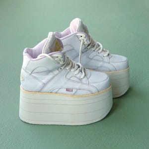 Op de afbeelding: Een paar witte platform sneakers met een high-top design. De schoenen hebben een dikke, gelaagde zool en witte veters. De binnenvoering is zacht lavendelkleurig. De sneakers zijn lichtblauw-grijs van kleur.