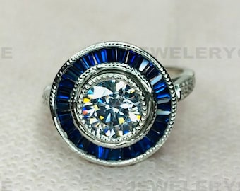 May include: A silver ring with a large, round, clear center stone surrounded by a halo of small, rectangular, deep blue stones. The ring has a detailed, vintage-inspired design.