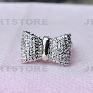 May include: A silver-colored bow-shaped charm, encrusted with small, sparkling stones. The charm has a polished, metallic finish and a central bar. The bow design is symmetrical and detailed, suitable for jewelry or accessories.