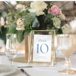 May include: A table number 10 card in a gold frame, with the word "table" in script and the number 10 in blue. The card also includes the names "Emily & Kevin" and the date "September 17, 2026". A floral arrangement and glassware are in the background.