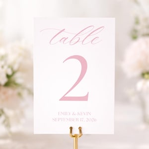 May include: A white table number card with the word "table" and the number "2" in pink script. The card also includes the names "Emily & Kevin" and the date "September 17, 2026". The card is held by a gold-colored metal clip.