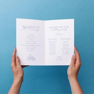 May include: A white wedding program with blue text, held open against a blue background. The program includes the words "Welcome to the Ceremony" and "Joined by our Wedding Party." The program lists the order of events and names of the wedding party.