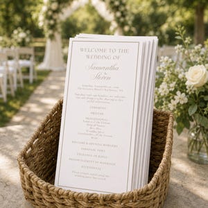 Puede incluir: Una pila de programas de boda blancos con bordes dorados y el texto Welcome to the Wedding of Samantha & Steven, Saturday, September 26, 2026, The Crescent Hotel, Rochester, NY, colocados dentro de una cesta de mimbre marrón.