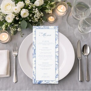 May include: A formal table setting with a white plate, silverware, and a menu card. The menu card has a blue floral design and the words "The Menu" at the top. White flowers and candles are arranged around the plate.
