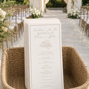Può includere: Una pila di programmi di nozze bianchi con bordi dorati e un elegante testo in corsivo che recita Welcome To The Wedding Of Emma & Tyler, con un'illustrazione floreale e l'ordine della cerimonia, posti all'interno di un cesto di vimini.