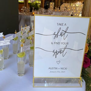 Op de afbeelding: Een goudkleurig bord met de tekst "TAKE A shot & FIND YOUR spot" en de namen "AUSTIN + NICKI" met de datum "31 januari 2025". Shotglaasjes met limoenpartjes en naambordjes staan op een witte tafel.