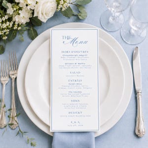May include: A rectangular white menu card with blue text and a thin blue border, centered on a white dinner plate. The menu lists courses including Hors d'oeuvres, Salad, Entrees, Sides, and Dessert, with the initials E & K and July 5th, 2026.