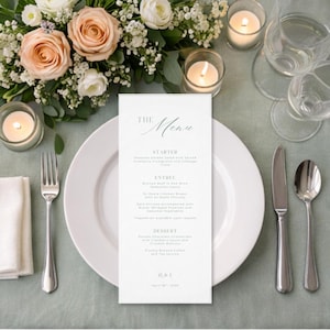 Op de afbeelding: Een bruilofttafelsetting met een wit menukaart, bord en bestek. De menukaart toont "The Menu" en de gangen. Perzik- en witte bloemen en kaarsen creëren een elegante sfeer.