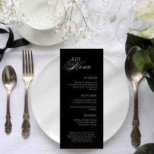 Könnte beinhalten: Elegante Tischdekoration mit einer schwarzen Menükarte. Das Menü, in weißer Schrift, beschreibt ein Drei-Gänge-Menü. Weiße Teller, Besteck und Blumenschmuck schaffen eine elegante Atmosphäre.