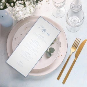 May include: A formal menu card with a pale blue border and elegant script. The menu is on a white plate with gold cutlery, wine glasses, and floral accents. The menu includes the text "The Menu" and a list of courses.